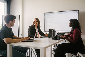 Professorin sitzt mit Studierenden an einem Besprechungstisch, im Hintergrund ein Bildschirm mit Programmiercode