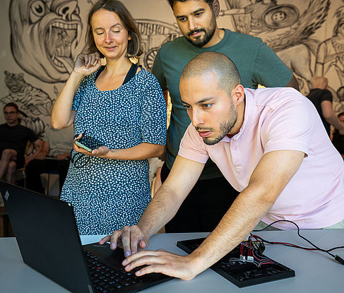 Drei Personen stehen um einen Laptop, wobei eine Person fokussiert daran arbeitet und die anderen interessiert zusehen.